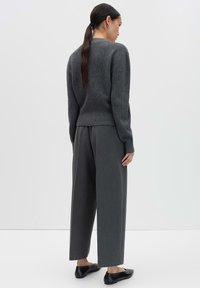 Donna con lunghi capelli scuri raccolti in una coda di cavallo, vestita con un maglione grigio a maglia, pantaloni larghi grigi e ballerine nere, in piedi di fronte a uno sfondo bianco.