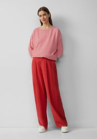 Rot gestreiftes Langarmoberteil mit weitem Ausschnitt, kombiniert mit lockeren roten Hosen und weißen Schuhen. Sanfte Texturen und lockere Passform.