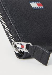 Black leather wallet featuring a silver zipper, embossed logo tag, and textured surface. The zipper's puller displays "TOMMY JEANS" branding.
