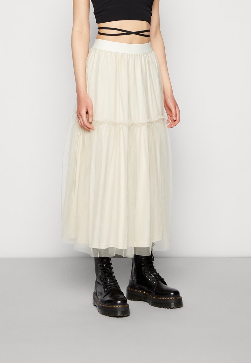 Personne portant une jupe maxi en tulle superposée couleur crème, un haut court noir avec des bretelles croisées et des bottes noires épaisses à lacets.