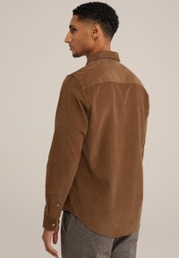 Camisa de pana marrón con cuello, mangas largas y botones en los puños; tejido texturizado, dobladillo ligeramente curvado en la parte trasera.