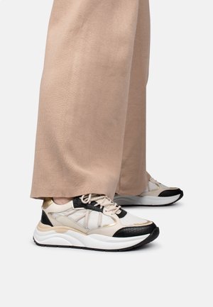 Personne portant un pantalon beige à jambes larges et des baskets blanches avec des accents noirs et dorés, debout sur un fond blanc.