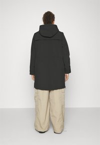 Veste noire avec capuche, ayant une coupe droite et un rabat à l'arrière. Associée à un pantalon cargo beige, ample.