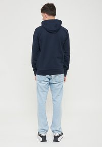 Felpa con cappuccio blu navy e polsini a coste, abbinata a jeans azzurri chiari. L'abbigliamento presenta una vestibilità rilassata e tasche posteriori standard.