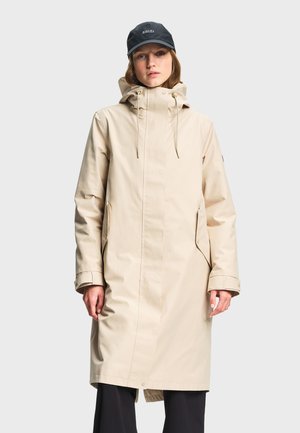 Femme portant un long imperméable à capuche beige et une casquette noire, debout devant un fond gris uni.