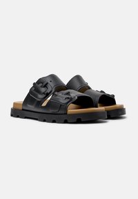 Black sandals with a wide strap, featuring prominent buckles, textured finish, and a tan-colored sole with a rugged pattern.