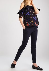 Schwarzes Off-Shoulder-Oberteil mit floralem Druck in Rot, Blau und Grün. Kombiniert mit navyblauen, schlank geschnittenen Hosen und schwarzen Slippern.
