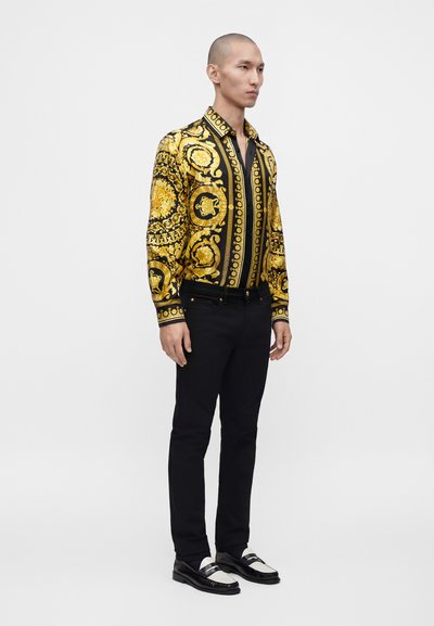 Homme debout portant une chemise à manches longues noire et dorée à motif, un pantalon noir et des mocassins noirs et blancs, légèrement tourné vers la droite.