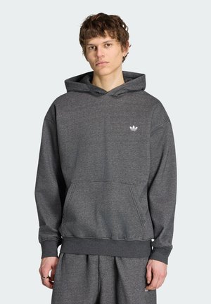Junger Mann trägt dunkelgrauen Adidas Hoodie mit Fronttasche und kleinem weißem Logo, kombiniert mit passenden Jogginghosen, steht vor schlichtem Hintergrund.