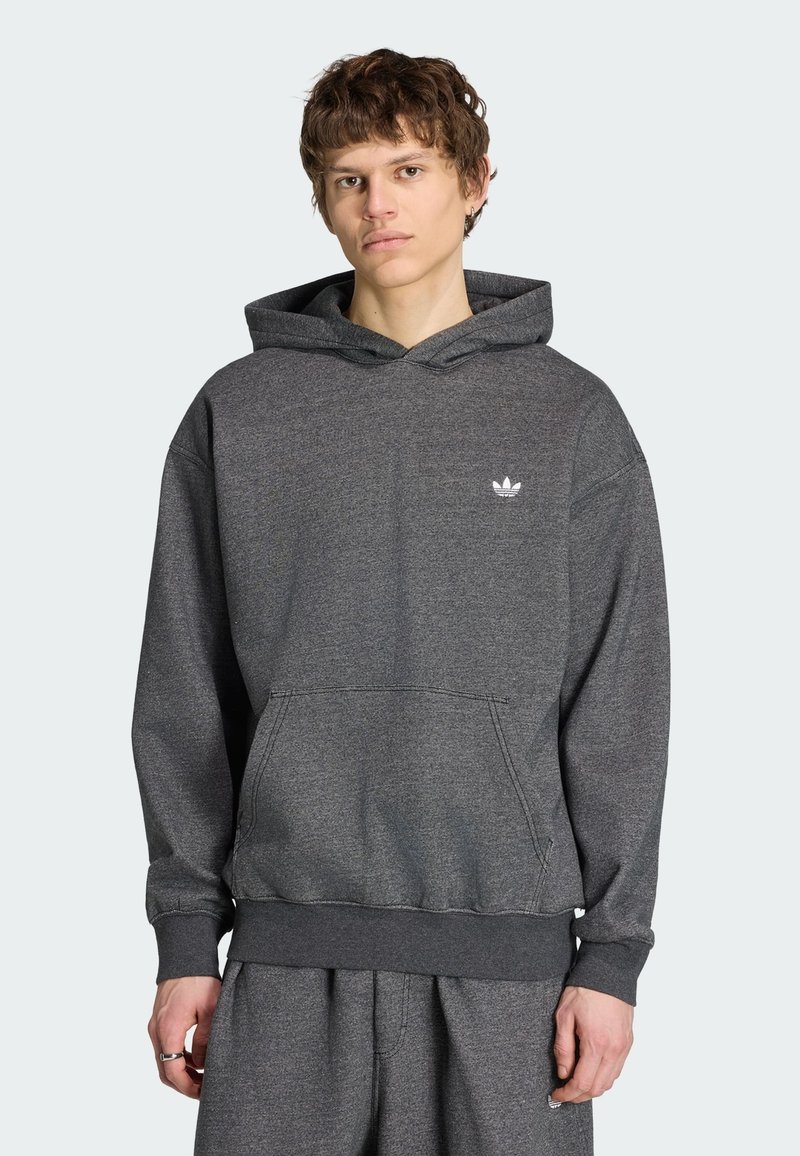 Jeune homme portant un sweat à capuche Adidas gris foncé avec une poche avant et un petit logo blanc, associé à un pantalon de jogging assorti, debout devant un fond uni.