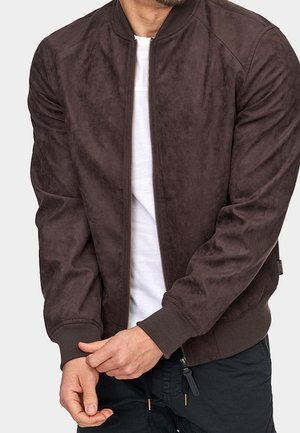 Homme portant une veste zippée en daim marron foncé par-dessus une chemise blanche, ajustant sa manche gauche avec sa main droite.