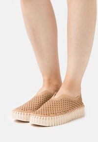 Beżowe buty slip-on z perforowaną tkaniną na górze i teksturowaną gumową podeszwą, z falbaniastymi krawędziami i niskim profilem.