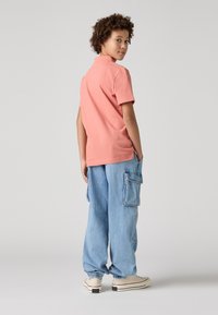 Camisa polo de algodón rosa de manga corta, combinada con pantalones cargo azul claro con bolsillos, y zapatillas beige con detalles negros.