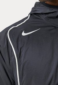Nike vindjakke i mørk navy, fremstillet af et let, struktureret stof. Inkluderer hvide pipede accenter og et fremtrædende hvidt Swoosh-logo.