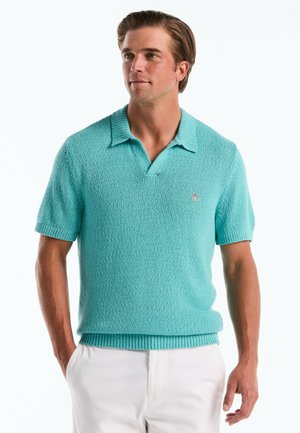 Mann trägt ein türkisfarbenes, kurzärmeliges Strick-Poloshirt mit geripptem Kragen und weiße Hose, steht mit den Händen in den Taschen.