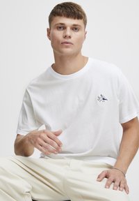 Hvid bomulds T-shirt med kortærmer, der har et lille broderet design på brystet. Glat tekstur, rund halsudskæring, afslappet pasform.