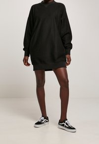 Abito oversized nero a coste con maniche lunghe e scollo a V, abbinato a sneakers in bianco e nero con lacci. Texture liscia, stile casual.