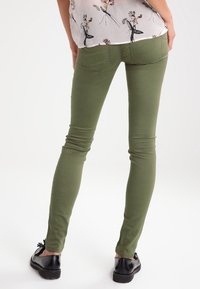 Pantalones vaqueros ajustados en verde oliva con una textura suave y bolsillos traseros estándar, combinados con zapatos planos negros y una blusa de patrón floral.