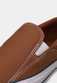 Chaussure en toile marron à enfiler avec coutures blanches et panneau latéral élastique blanc sur une semelle blanche.