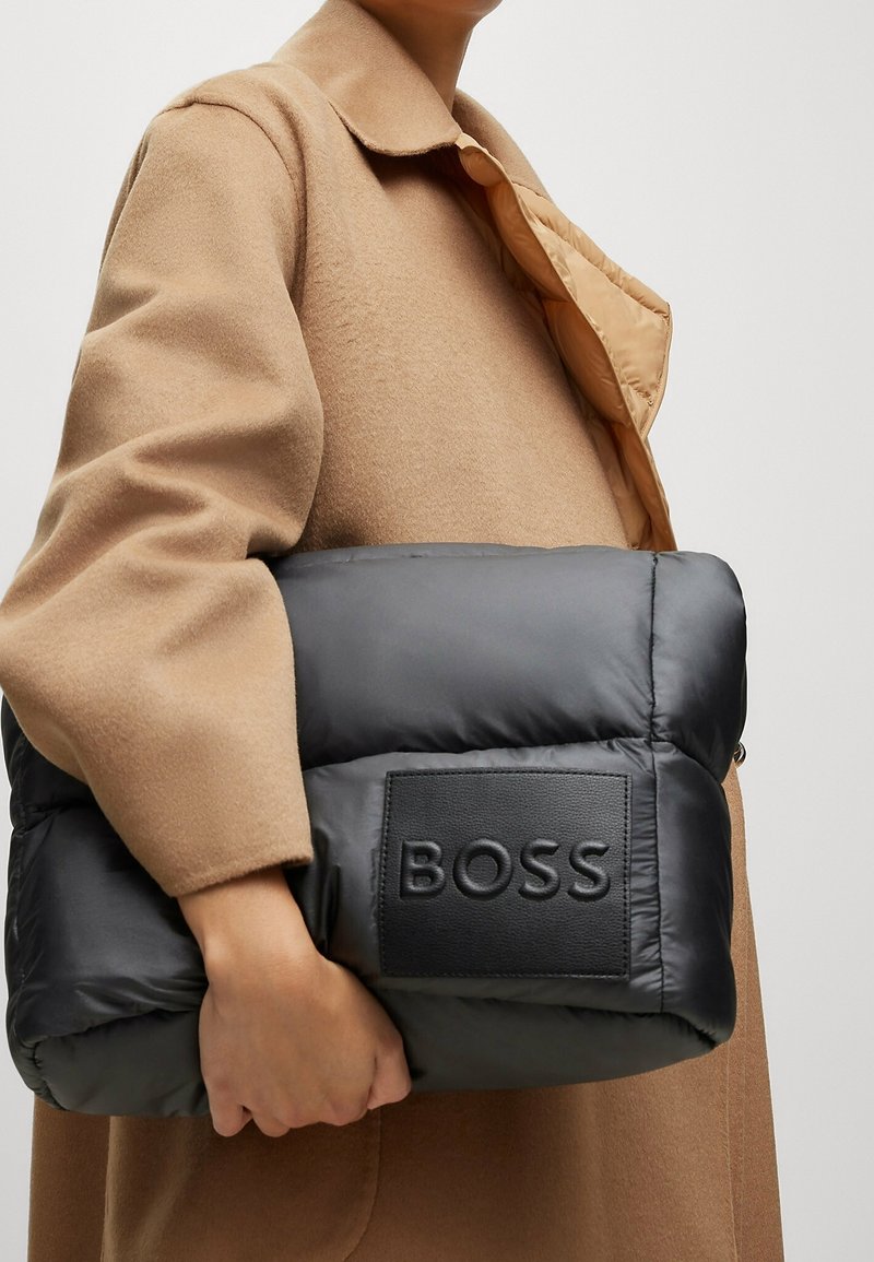 Personne portant un manteau camel tenant un grand sac noir matelassé avec un patch logo BOSS visible.
