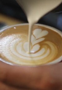Un art latte dans une tasse blanche présente un design de mousse brun clair avec un motif de cœur et de spirale sur la surface crémeuse du café. Une main tient la tasse.