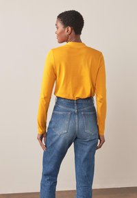 Langærmet gul bluse kombineret med højtaljede blå jeans. Blusen har et simpelt design, mens jeansene har en klassisk denimtekstur.