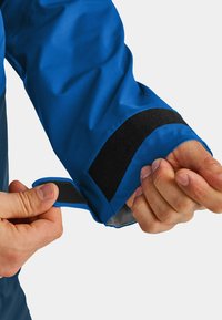 Blaue wasserdichte Jacke mit einem Klettverschluss am Ärmel, während Hände den Ärmel ajustieren, und die strukturierte Innenausstattung sichtbar ist.