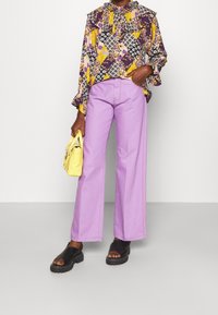 Pantalon large violet avec des poches avant, associé à un chemisier à motifs avec des volants. Tient un petit sac jaune et porte des sandales noires.