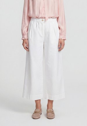 Frau trägt weiße, weit geschnittene, gekürzte Hosen, eine hellrosa Langarmbluse und beige Loafers mit goldener Kettendetail.