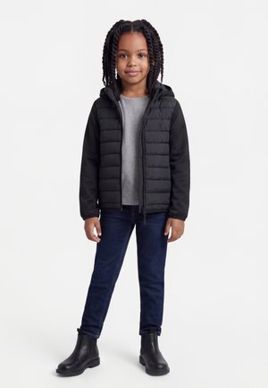 Νεαρή κοπέλα με πλεγμένα μαλλιά, φοράει μαύρο φουσκωτό μπουφάν, γκρι μπλούζα, σκούρο τζιν και μαύρες μπότες, στέκεται μπροστά σε λευκό φόντο.