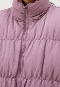 Tjock väst i ljus lavendel med hög krage, centrerad dragkedja och strukturerat tyg med horisontell sömnad för extra detalj.