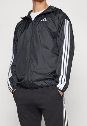 Man draagt een zwarte Adidas trainingsjas met witte strepen op de mouwen en bijpassende zwarte broek met witte strepen aan de zijkant, handen in de zakken.