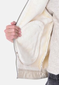 Giacca beige con un morbido rivestimento in pile color crema. Chiusura a zip e tasche interne visibili. Il tessuto ha una texture liscia.