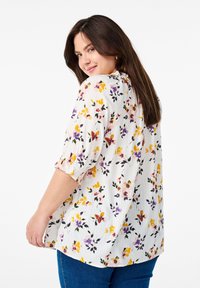Blusa con motivo floreale in tessuto leggero e testurizzato con maniche elastiche. Presenta una base bianca con fiori viola, gialli e arancioni.