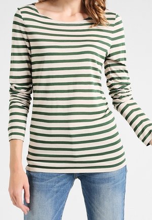 Femme portant un t-shirt à manches longues rayé horizontalement vert et blanc, associé à un jean bleu clair, debout devant un fond uni.