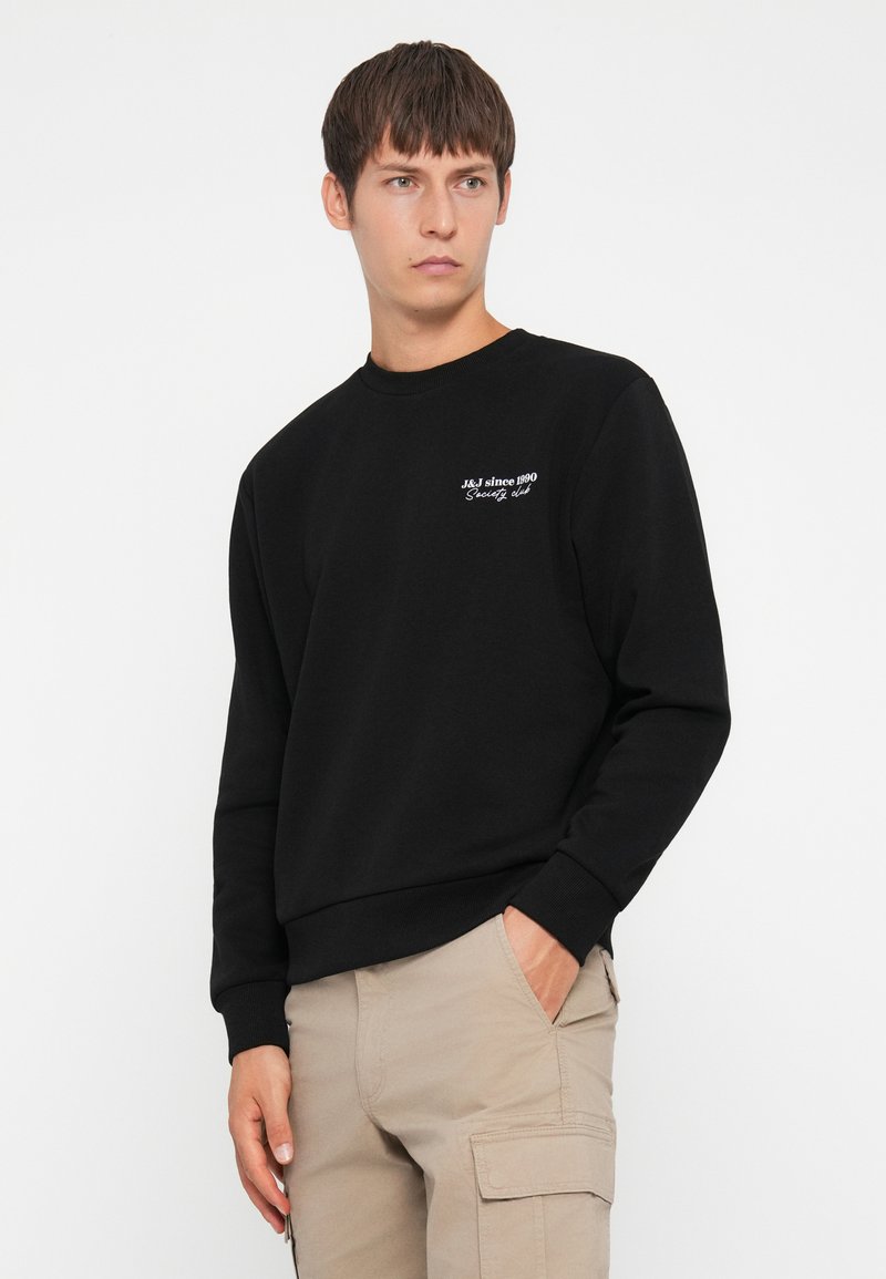 Sweatshirt noir à manches longues, col rond côtelé et texte brodé à l'avant. Associé à un pantalon cargo beige. Texture de tissu lisse.