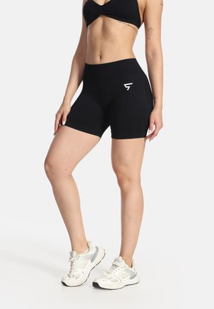 Frau trägt schwarze, hoch taillierte Sportshorts und einen schwarzen Sport-BH, kombiniert mit weißen Laufschuhen, vor einem einfachen Hintergrund stehend.
