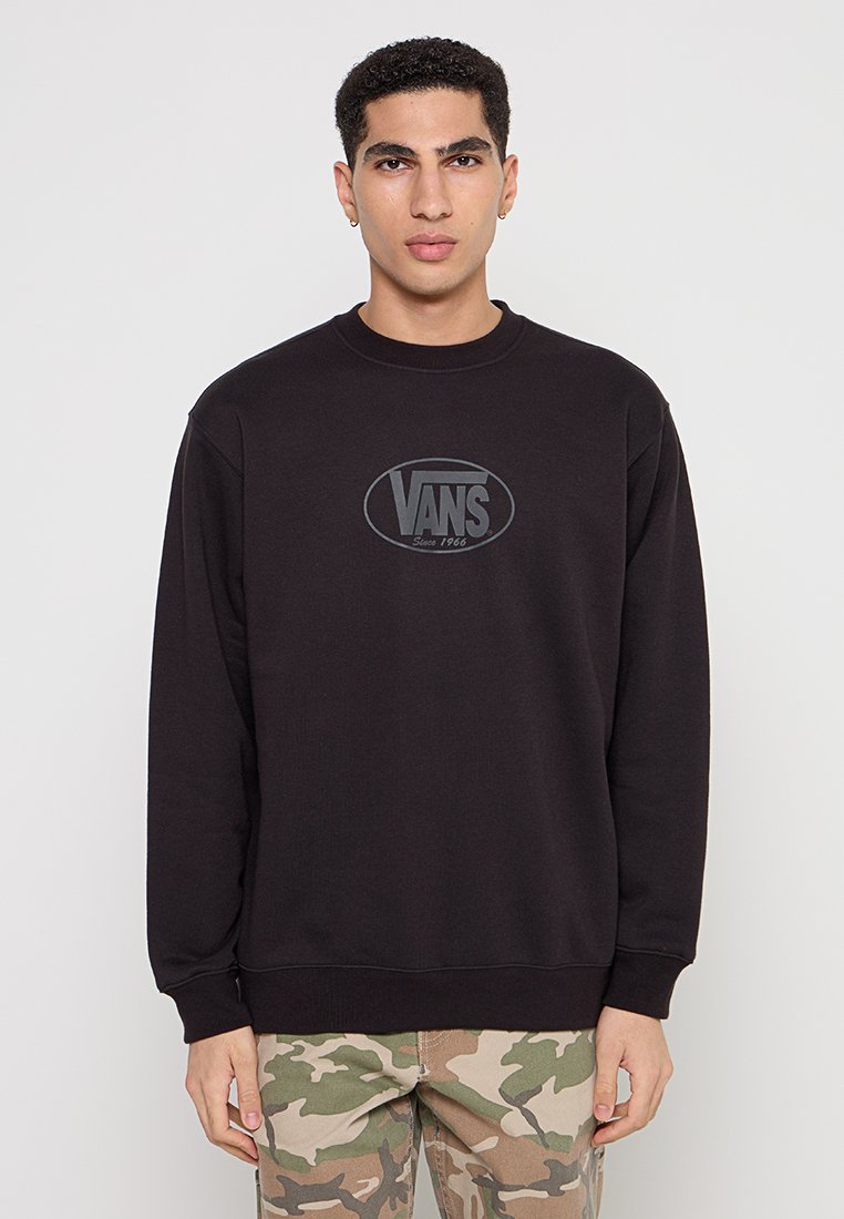Felpa nera con logo ovale testurizzato con il testo "VANS", collo a giro e polsini a costine. Indossata con pantaloni a fantasia mimetica.