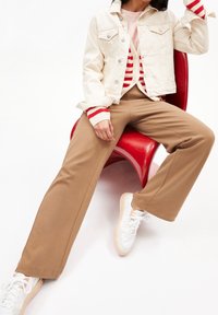 Veste en denim ivoire, pantalon large beige, pull rayé rouge et blanc, et baskets blanches avec semelles beiges, assis sur une chaise rouge.