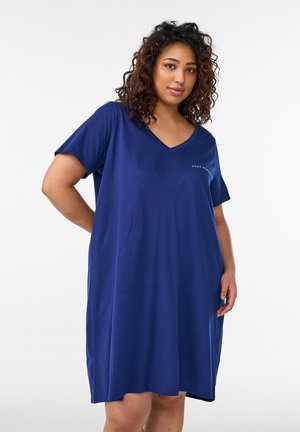 Donna con capelli ricci che indossa un abito blu largo a maniche corte con scollo a V e testo sottile sul petto.