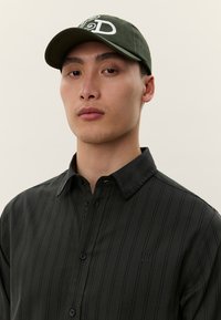 Νεαρός άνδρας με ουδέτερη έκφραση, ντυμένος με σκούρο πράσινο πουκάμισο με ρίγες και σκούφο ελιάς με λευκά γράμματα.