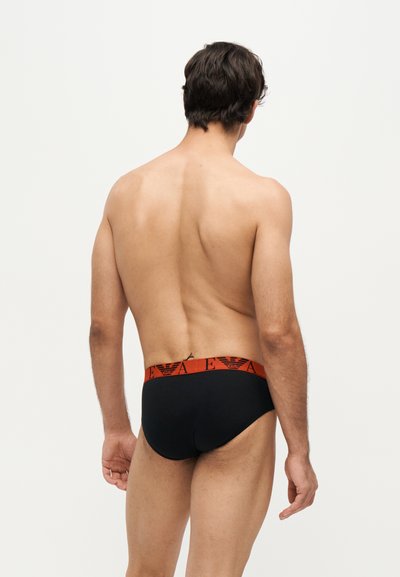 Homme aux cheveux courts et foncés portant un slip noir avec une ceinture rouge affichant un logo, debout dos au spectateur sur un fond clair uni.