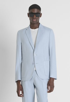 Costume bleu clair au design ajusté, fabriqué en tissu texturé ; fermeture à un bouton, deux poches avant, associé à un haut en maille blanche.