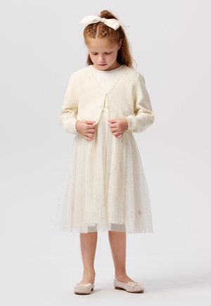 Helle cremefarbene Strickjacke mit Knopfverschluss über einem halbtransparenten Tüllkleid, das subtile Funken enthält. Abgerundet mit weichen Ballettschuhen.