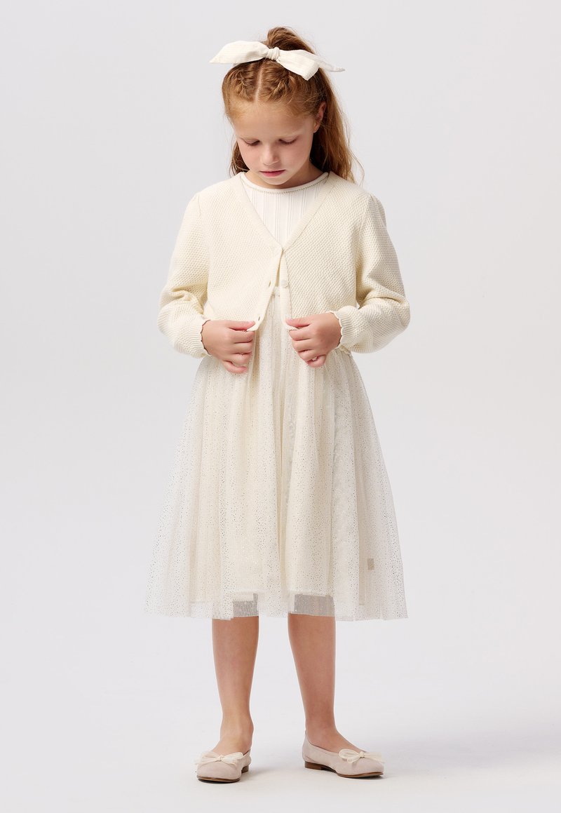 Cardigan léger crème avec fermeture à boutons sur une robe en tulle semi-transparente ornée de subtils paillettes. Complété par des ballerines douces.