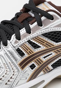 Gros plan d’une basket Asics noire et marron avec des superpositions argentées, tissu en mesh, lacets noirs et semelle blanche texturée.