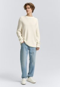 Jong persoon in een crèmekleurige oversized trui, lichtblauwe rechte jeans en witte sneakers, staande met één hand in de zak.