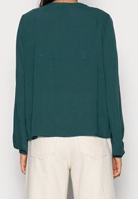 Blouse turquoise à manches longues, col rond et coupe ample, associée à un pantalon taille haute de couleur claire. Tissu à texture lisse.