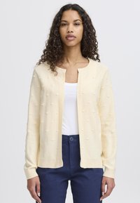 Světle žlutý cardigan s texturovanými bobble detaily, žebrovanými manžetami a lemem, nošený přes bílé tričko, v kombinaci s tmavě modrými kalhotami.