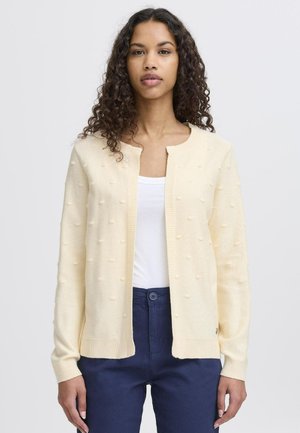 Světle žlutý cardigan s texturovanými bobble detaily, žebrovanými manžetami a lemem, nošený přes bílé tričko, v kombinaci s tmavě modrými kalhotami.